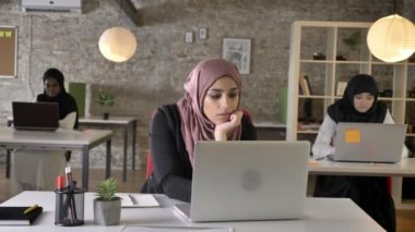 Üç genç Müslüman kadının içinde oturan ve esneme ve çalışma, Müslüman kadının modern ofiste dizüstü bilgisayarda yazarak hijab yorgun