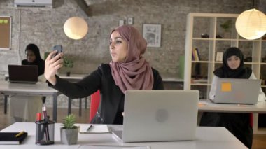 Üç genç Müslüman kadının içinde oturan ve modern ofis, büyüleyici kadın işçi selfie telefon ile alarak çalışan hijab gülümseyen