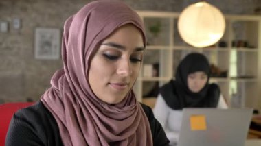 Çalışma, kamera seyir ve gülümseyen, oturma ve modern ofis dizüstü bilgisayarda yazarak iki Bayan Tesettür genç Müslüman kadın portresi