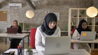 Üç genç Müslüman kadın oturuyor ve modern ofis, laptop, kameraya gülümseyen yazarak islam kadın işçi çalışan hijab içinde