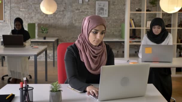 Tres mujeres musulmanas jóvenes en hijab sentado y escribiendo en el ordenador portátil en la oficina moderna, mujer negra en el trabajo de hijab — Vídeo de stock