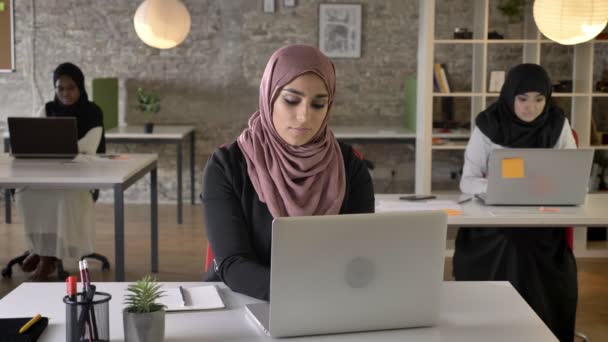 Tres jóvenes musulmanas en hijab sentadas y trabajando en la oficina moderna, musulmana hermosa mujer mirando a cámara y sonriendo — Vídeos de Stock