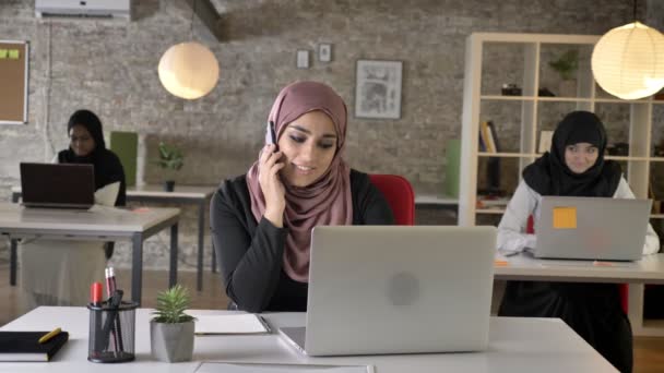 Tres jóvenes musulmanas en hijab sentadas y trabajando en una oficina moderna, hermosa mujer musulmana hablando por teléfono, sonriendo — Vídeos de Stock