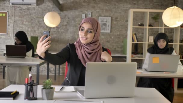 Три молодих мусульманських жінки в хіджабі, сидячи і працюючи в сучасному офісі, досить мусульманська жінка бере селфі з телефоном, посміхаючись — стокове відео