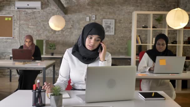 Három fiatal muzulmán nők a hidzsáb ül és a modern iroda, szép muszlim nő beszél telefonon, mosolyogva — Stock videók