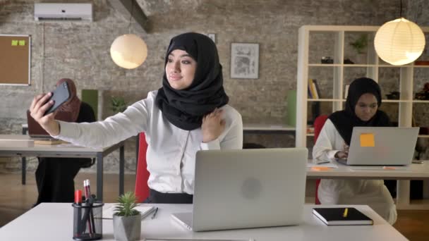Three young muslim womens in hijab sitting and typing on laptop in modern office, charming muslim women taking selfie with phone — Stock Video
