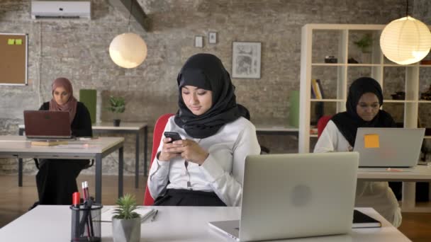 Drie jonge islamitische womens in hijab zitten en typen op laptop in moderne kantoor, charmante werkneemster texting op telefoon — Stockvideo