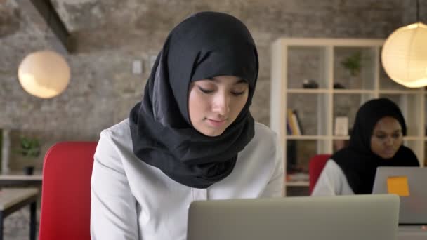 Πορτρέτο των νέων μουσουλμάνων γυναικών στο χιτζάμπ εργασίας και ψάχνει στην κάμερα, χαμογελώντας, δύο γυναικών που κάθεται στο σύγχρονο γραφείο — Αρχείο Βίντεο