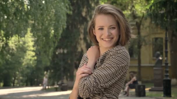 Joven hermosa chica feliz está de pie y sonriendo en el parque durante el día en verano, mirando a la cámara — Vídeos de Stock