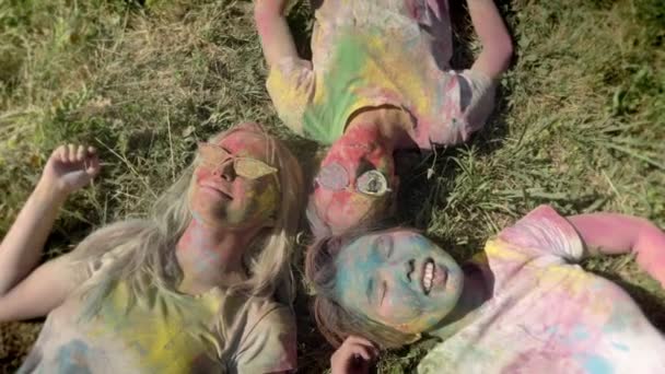 Tres jóvenes amigas multiétnicas felices están tumbadas en la hierba en el festival holi durante el día en verano, concepto de amistad, concepto de vacaciones — Vídeo de stock