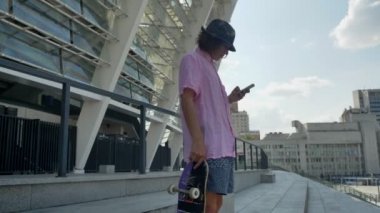 Genç adam hipster mesaj yazarak smartphone, kaykay elinde tutan, kentsel kavramı, iletişim kavramı, yan görünüm