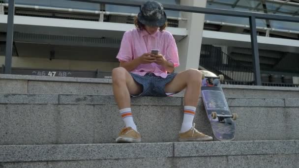 Молодой человек хипстер печатает сообщение на смартфоне, скейтборде, городской концепции, коммуникационной концепции, Dolly shot — стоковое видео