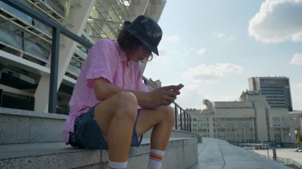 Young man hipster is typing message on smartphone, legs on skateboard, urban concept, communication concept, side view — Stock Video