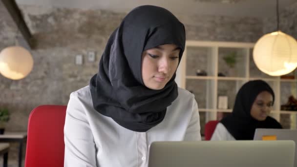 笑みを浮かべて つの女性 それらの つのオフィスに座って作業とカメラに探してヒジャーブでの若いイスラム教の女性黒のヒジャーブの女性 — ストック動画