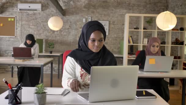 Zwarte jonge moslimvrouw in hijab typen op laptop en schrijven op papier, drie prachtige islamitische womens werkzaam in kantoor — Stockvideo
