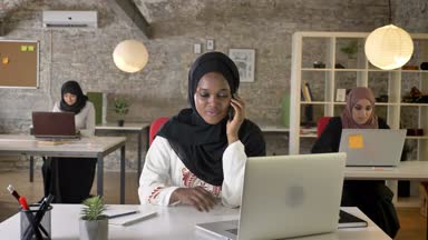 Modern ofis, telefon oturma ve dizüstü bilgisayarda çalışan üç oldukça Müslüman kadın konuşurken hijab genç siyahi Müslüman kadınları
