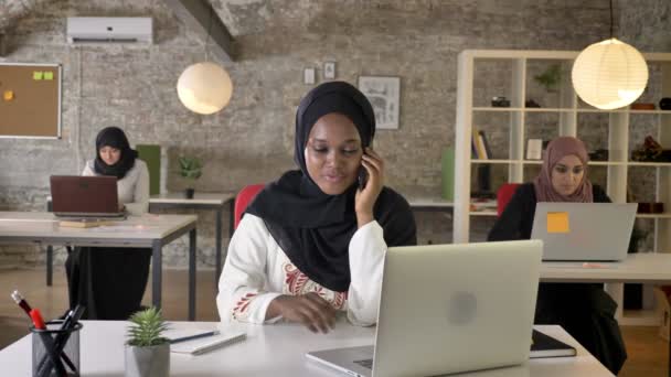 Jóvenes mujeres musulmanas negras en hijab hablando por teléfono en la oficina moderna, tres mujeres musulmanas bastante sentado y trabajando en el ordenador portátil — Vídeos de Stock