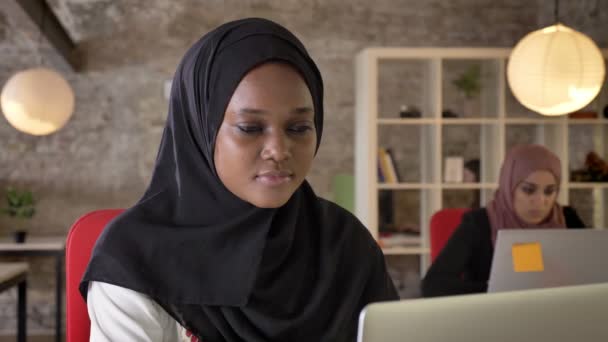 Портрет молодых черных мусульманских женщин в хиджабе, работающих на ноутбуке, сконцентрированных, две очаровательные женщины, сидящие в современном офисе — стоковое видео