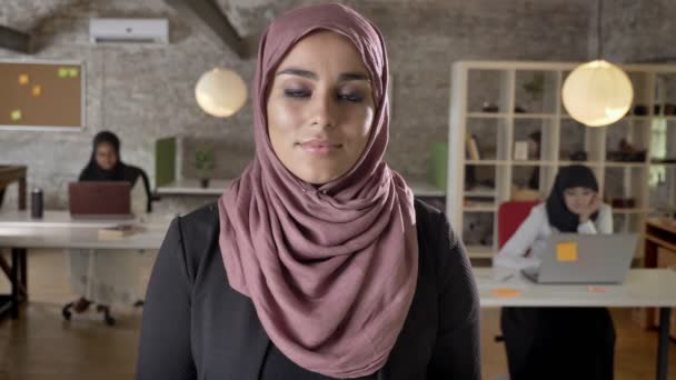 Portrait of young muslim women in hijab looking in camera, two womens sitting and typing on laptop in office at background — Stok Video