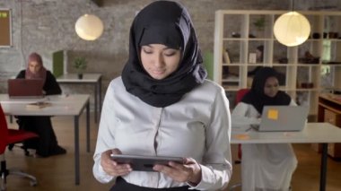 Tablet kullanma, kamera seyir, gülümseyen, laptop modern ofisindeki tipik iş Bayan Tesettür genç Müslüman kadınları