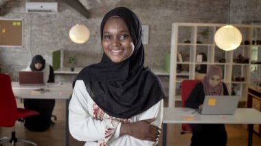 Genç Müslüman kadın kamera seyir, gülümseyen, üç kadın laptop modern arka plan ofisinde çalışan hijab siyah