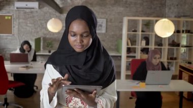 Tablet kullanma, kamera seyir, gülümseyen, modern ofis dizüstü bilgisayarda yazarak İslam iş Bayan Tesettür genç siyahi Müslüman kadınları