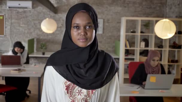 Wanita muslim muda afrika dalam hijab berdiri dan melihat ke kamera, prihatin, tiga wanita bekerja pada laptop di kantor modern — Stok Video