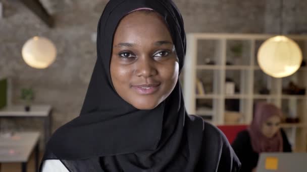 Портрет молодых черных мусульманских женщин в хиджабе, смотрящих в камеру, улыбающихся, женщин, работающих на ноутбуке в современном офисе в задней части — стоковое видео
