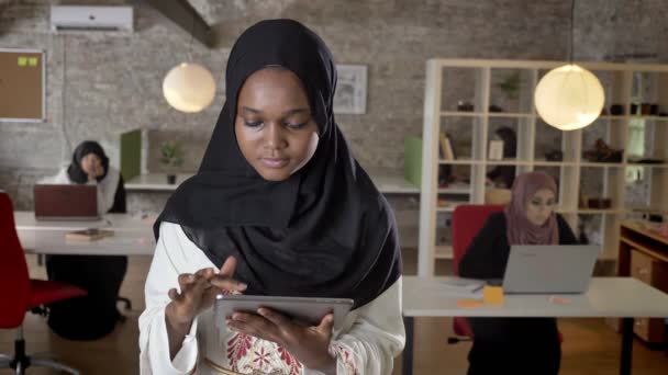 Junge schwarze muslimische Frauen im Hijab mit Tablet, in die Kamera guckend, lächelnd, islamische Geschäftsfrauen tippen auf Laptop im modernen Büro — Stockvideo