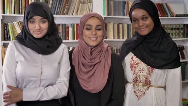 Три молодые мусульманские женщины в хиджабе стоят в библиотеке и смотрят в камеру, улыбаясь и радуясь — стоковое видео