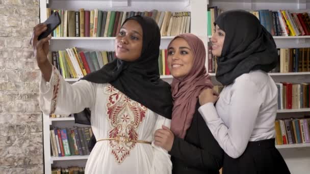 Три молодые симпатичные мусульманские женщины в хиджабе делают селфи и стоят в библиотеке, друзья фотографируют, улыбаются и счастливы — стоковое видео