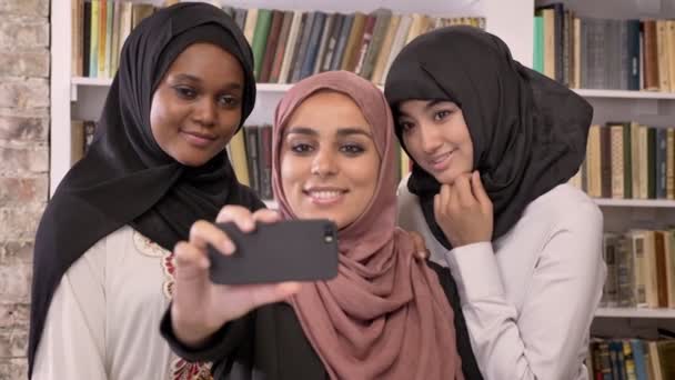 Три молодые красивые мусульманские женщины в хиджабе делают селфи в библиотеке и смотрят на телефон после, улыбаясь — стоковое видео