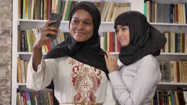 Jeunes femmes musulmanes noires en hijab avec un ami blanc prenant selfie dans la bibliothèque, souriant et heureux, étudiants islamiques — Video