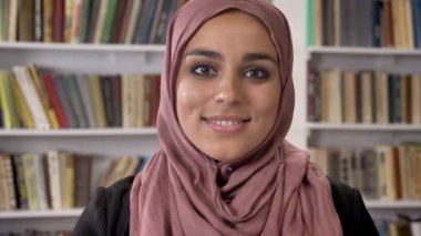 Genç Müslüman kadınları kamera seyir ve gülümseyerek, kitaplıkta ayakta hijab portresi