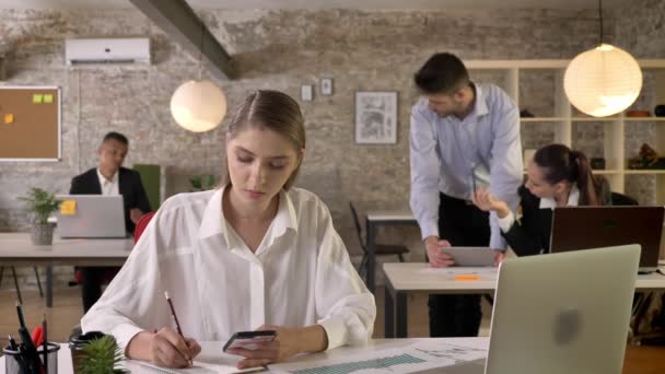 Young busy businesswoman is working in office, writing down, holding smartphone in hand, discussion on background, working concept, business concept, communication concept — Stock Video