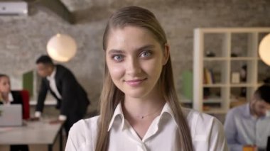Genç çekici iş kadını ofisteki kameraya bakıyor, gülümsüyor, meslektaşları teknolojilerle, çalışma konseptiyle, iş konseptiyle, bulanık geçmişiyle iletişim kuruyor.