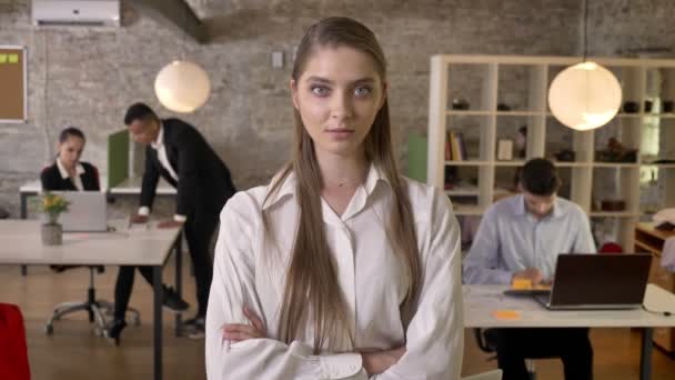 Jeune femme d'affaires charmante regarde à la caméra dans le bureau, bras croisés, ses collègues sont en réseau avec les technologies, concept de travail, concept d'entreprise, arrière-plan flou — Video