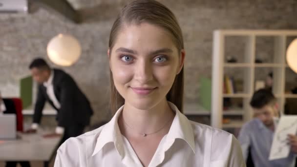 Jonge mooie zakenvrouw is kijken op camera in kantoor, glimlachen, haar collega's zijn netwerken met onscherpe achtergrond, technologieën, werk concept, bedrijfsconcept. — Stockvideo