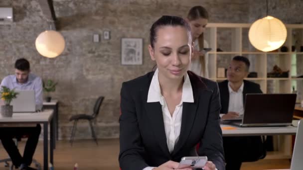 Giovane donna d'affari sta leggendo e digitando il messaggio sullo smartphone in carica, i suoi colleghi sono in rete con le tecnologie, concetto di lavoro, concetto di business, concetto di comunicazione — Video Stock