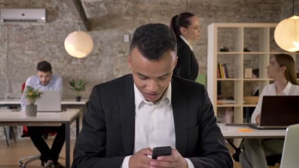 Giovane imprenditore afro-americano sta digitando il messaggio sullo smartphone in carica, i suoi colleghi sono in rete con le tecnologie, concetto di lavoro, concetto di business, concetto di comunicazione — Video Stock