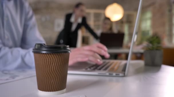 Cup of coffee on table in office, businessman is typing on laptop, his colleagues are networking with technologies, blurred background, working concept — Stock Video