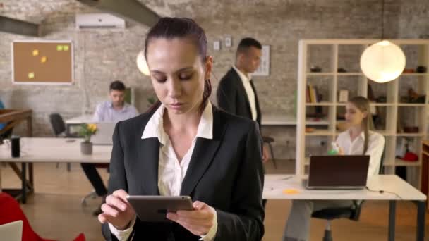 Giovane affascinante donna d'affari sta toccando tablet in ufficio, guardando alla telecamera, i suoi colleghi sono in rete con le tecnologie, concetto di lavoro, concetto di business, concetto di comunicazione — Video Stock