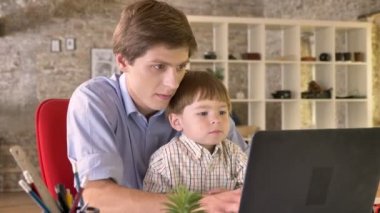 Küçük oğlu tutarak ve dizüstü bilgisayar, meşgul, çalışan genç baba modern ofisinde oturuyor