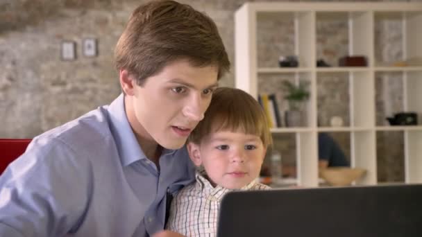 Padre joven sosteniendo a su hijo sonriente y mirando a la computadora portátil, sentado en la oficina moderna, feliz — Vídeos de Stock