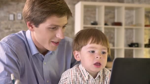 Junger Vater hält seinen kleinen Sohn und blickt auf Laptop, konzentriert und ernst, sitzt in modernem Büro — Stockvideo