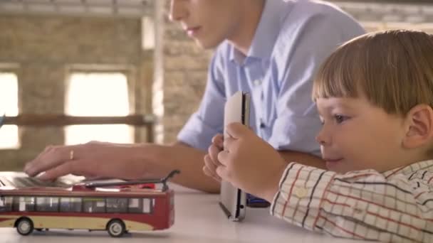 Pequeño hijo mirando la tableta cuando su padre joven que trabaja en el ordenador portátil, sentado en la oficina de ladrillo moderno — Vídeos de Stock