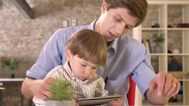 Jeune père regardant tablette avec son petit fils, enfant ennuyé par son père, assis dans un bureau moderne en brique — Video