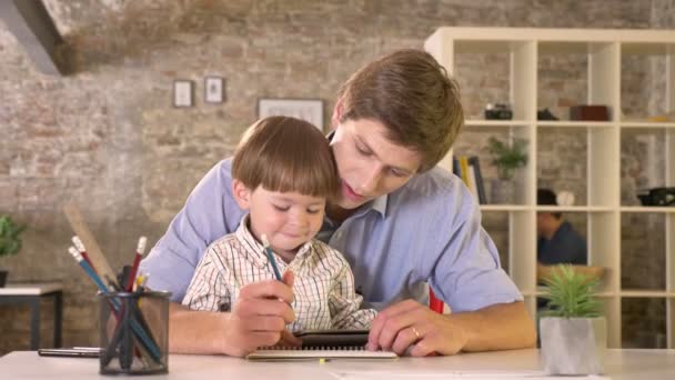 Joven padre sosteniendo a su pequeño hijo y escribiendo en papel, niño sosteniendo tableta, sentado en la oficina moderna — Vídeos de Stock