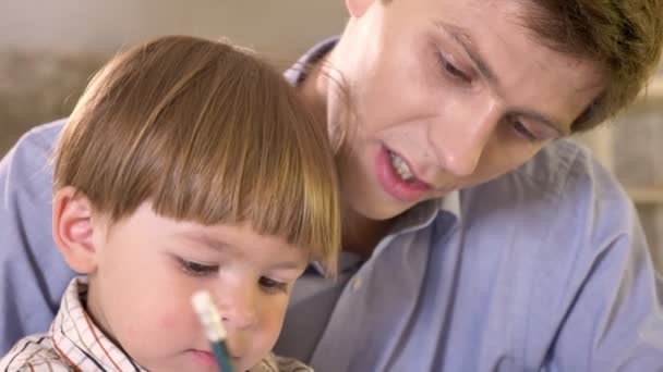 Fiatal apa és a fia kis festés a papír és a gazdaság tabletta, apa segít, és hogyan kell írni a fia oktatásának — Stock videók
