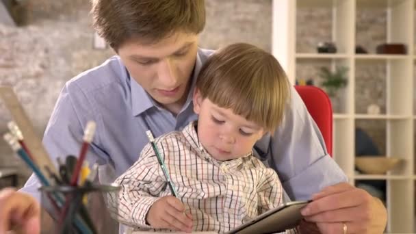Молодой отец держит своего маленького сына, сидит за столом и пишет на бумаге, современный офисный фон — стоковое видео